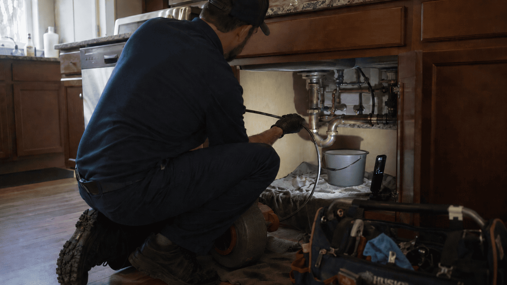 Professional plumber using a drain auger under a kitchen sink to clear a clogged drain line